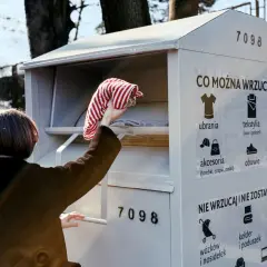 Kobieta wrzuca ubrania do szarego kontenera na odzież. Na kontenerze widnieje napis "CO MOŻNA WRZUCAĆ?".
