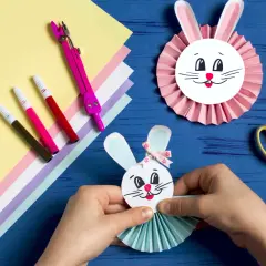 Ręce tworzą wielkanocnego zajączka z papieru, obok materiały i przybory plastyczne.
