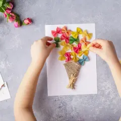Dziecko tworzy kolorowe laurki na Dzień Mamy, przyklejając makaron farfalle na kartce.