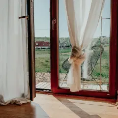 Białe zasłony przewiązane węzłem, jak skrócić zasłony, by wyglądały stylowo. Widok na zieloną łąkę i dom.