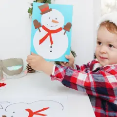 Dziecko w czapce Mikołaja pokazuje kartkę z bałwankiem, inspirując do zabawy: jak zrobić bałwanka.