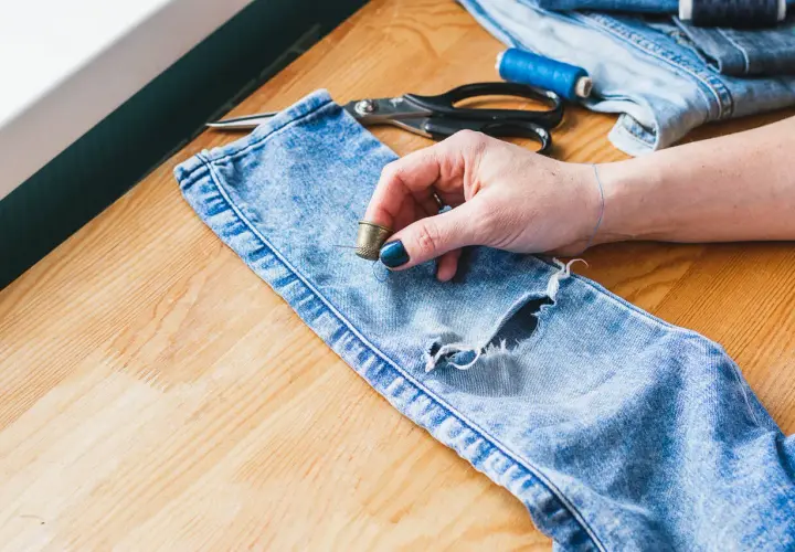 Ręczne szycie dziury w spodniach. Dłoń z naparstkiem i igłą przygotowuje się do naprawy jeansów. Obok leżą nożyczki i nici.