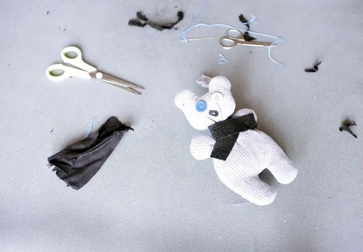 Uroczy kot ze skarpetki, z guzikami zamiast oczu, leży obok nożyczek i resztek materiału.