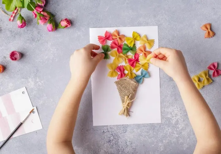 Dziecko tworzy kolorowe laurki na Dzień Mamy, przyklejając makaron farfalle na kartce.
