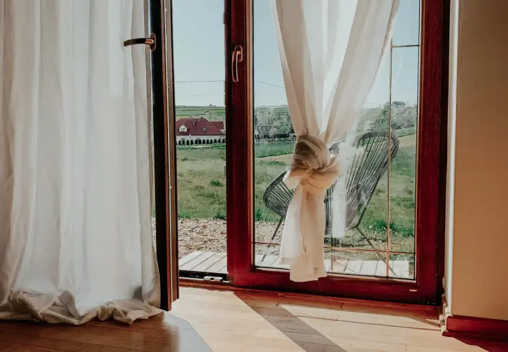 Białe zasłony przewiązane węzłem, jak skrócić zasłony, by wyglądały stylowo. Widok na zieloną łąkę i dom.