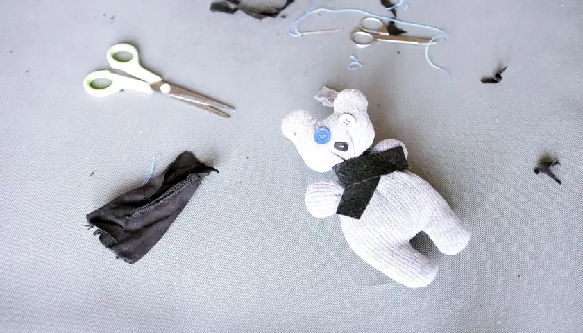 Uroczy kot ze skarpetki, z guzikami zamiast oczu, leży obok nożyczek i resztek materiału.