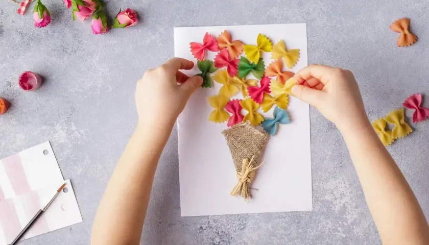 Dziecko tworzy kolorowe laurki na Dzień Mamy, przyklejając makaron farfalle na kartce.